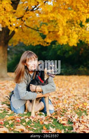 Fröhliche junge kaukasische Frau, die auf dem Boden sitzt und den Hund umarmt. Besitzer, der mit Haustier am Herbstfalltag spazieren geht. Beste Freunde, die im Freien Spaß haben. Freundschaft von h Stockfoto