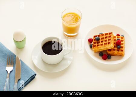 Blick aus dem Blickwinkel auf ein köstliches Gourmet-Frühstück mit süßen Waffeln, Ei und Orangensaft neben einer Tasse schwarzen Kaffee auf einem weißen Tisch, Kopierraum Stockfoto