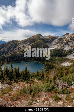 Ein Bergsee in einem Becken ist von der Seite eines Berges aus sichtbar, umgeben von westlichen Lärchen, unter einem blauen Himmel mit weißen und hellgrauen Wolken Stockfoto