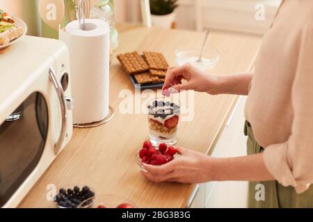 Warme getönte Nahaufnahme von unkenntlichen jungen Frau, die Joghurt Parfait mit frischen Beeren und Müsli während der Vorbereitung gesundes Frühstück am Morgen, c Stockfoto