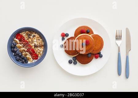 Draufsicht auf minimale Zusammensetzung von köstlichen goldenen Pfannkuchen mit frischen Beeren neben Granola Platte auf weißem Hintergrund, gesundes Frühstück dekoriert Stockfoto