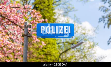 Zentrierte Ansicht auf einem blauen Polizei-Schild. Vor einer deutschen Polizeistation. Verschwommener Hintergrund mit blühenden Bäumen und blauem Himmel mit Wolken. Panorama. Stockfoto