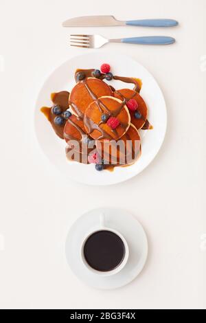 Draufsicht bei minimaler Zusammensetzung von köstlichen goldenen Pfannkuchen mit Schokolade und frischen Beeren neben Kaffeetasse auf weißem Hintergrund, Frühstück dekoriert Stockfoto