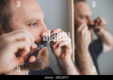 Selbstversorgung während der Quarantäneisolierung. Erwachsene gut aussehende kaukasischen Mann Schneiden Schnurrbart und Bart sich persönlich mit der Schere zu Hause. Coronavirus o Stockfoto