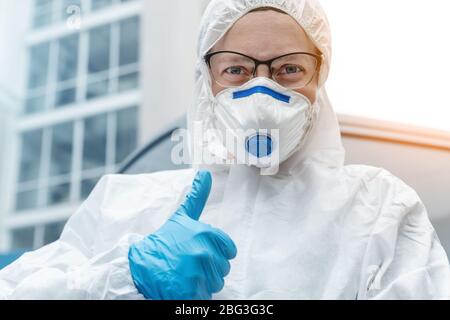 Porträt der müde erschöpft Ärztin, Wissenschaftlerin oder Krankenschwester trägt Gesichtsmaske und biologische Hasmat psa Anzug zeigt Vertrauen Daumen nach oben wie Stockfoto