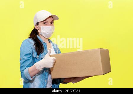Sichere Lieferung. Frauen-Kurierin in Schutzmaske und Handschuhe mit Karton-Box auf gelbem Hintergrund. Anti-Coronavirus-Konzept. Stockfoto