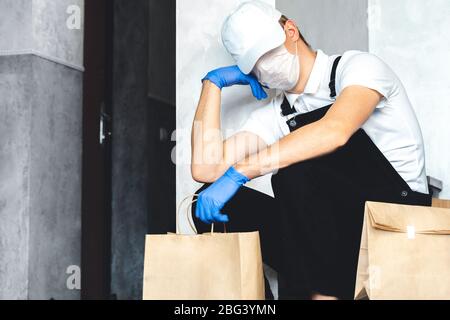 Gelangweilter Kurier in schützende chirurgische Maske und medizinische Handschuhe liefern Papiertüte mit Essen zum Mitnehmen. Lieferung Mann wartet an der Tür. Kontaktlos del Stockfoto