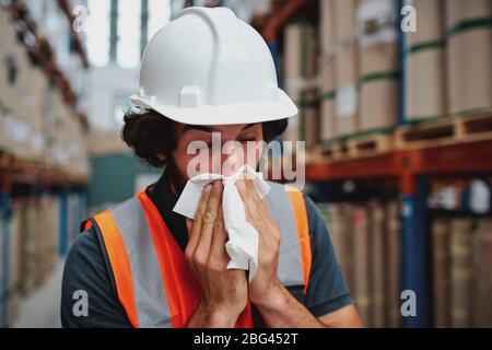 Junge Lagerarbeiter fühlen sich krank, während sie niesen und husten in der Fabrik, die in der Nähe von verpackten Kartons im Regal steht Stockfoto