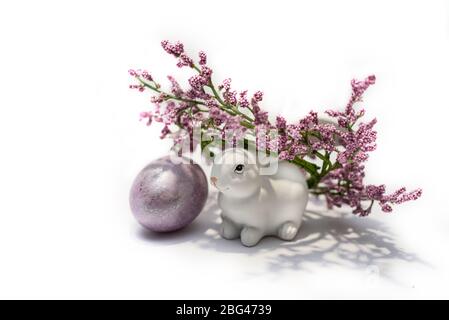 Ein weißes Hasen Kaninchen mit Kranz von lila Blüten und farbigen osterei in der Nähe. Konzept der Osterferien. Stockfoto