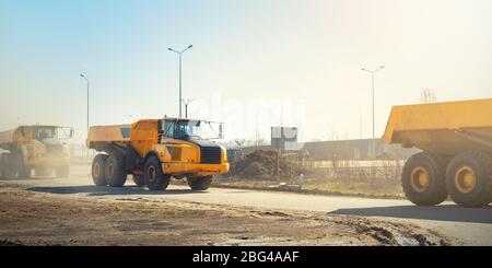 Viele große Gelenkwagen, die am sonnigen Tag mit blauem Himmel auf der neuen Straßenbaustelle fahren, sind mit gelben Müllhalden befahren Stockfoto