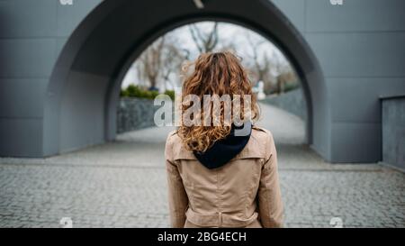 Rückansicht junge Frau mit lockigen Haaren in beige Mantel steht und schaut auf Stadtpark an düsteren Tag gekleidet. Stockfoto