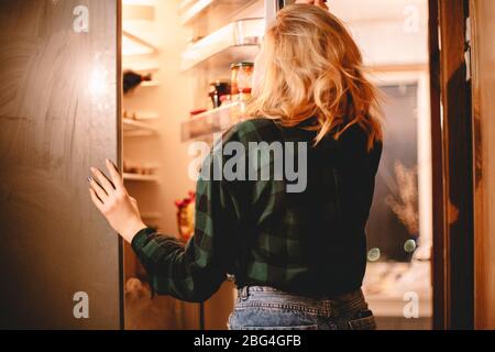 Junge hungrige Frau, die zu Hause im offenen Kühlschrank steht Stockfoto