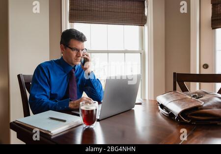 Mann auf dem Handy, der von zu Hause aus mit einem Computer am Esstisch arbeitet. Stockfoto