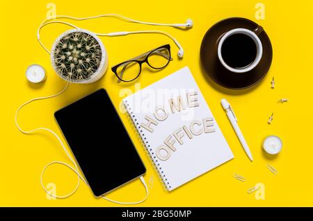 Flacher moderner Arbeitsplatz mit Tablet, Ohrhörer, Kaffeetasse, Notizblock, Stift, Brille und Kaktus. Briefe mit der Aussage „Home Office“. Draufsicht auf gelb b Stockfoto