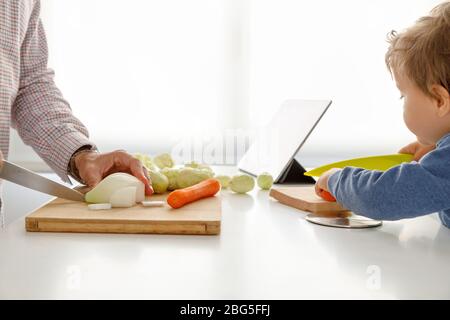 Ein kleiner Junge hilft seinem Vater, Gemüse mit einer Tablette im Hintergrund zu schneiden Stockfoto