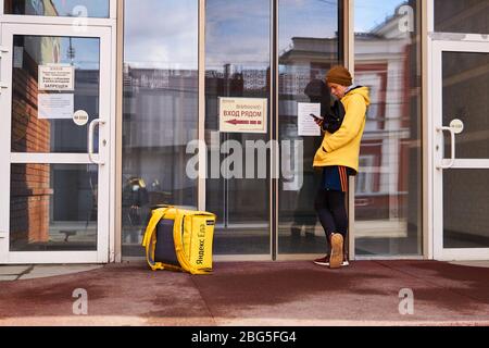 Perm, Russland - 19. April 2020: Fahrradkurier eines Online-Lebensmittelzustelldienstes ruft einen Kunden während der Quarantäne in C vor einer geschlossenen Tür an Stockfoto