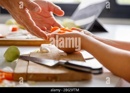 Ein Mädchen gibt ihrem Vater eine Handvoll gehacktes Gemüse, während sie zusammen mit einer Tablette im Hintergrund kochen Stockfoto