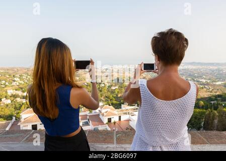 Rückansicht von zwei Touristen-Frauen, die mit dem Telefon Fotos von der wunderschönen Landschaft des Dorfes Mijas in Spanien machen Stockfoto