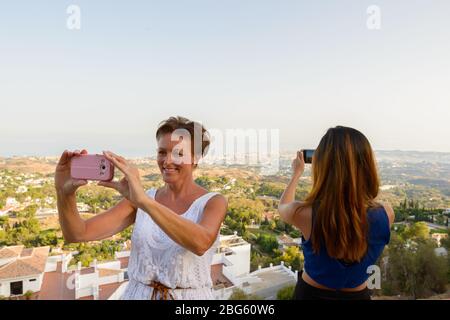 Zwei glückliche Touristen Frauen, die mit dem Telefon Fotos von der wunderschönen Landschaft des Dorfes Mijas in Spanien machen Stockfoto