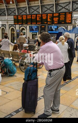 LONDON, ENGLAND - JULI 2018: Mann, der sich auf einem Koffer auf der Bahnhofshalle am Bahnhof Paddington stützt, beobachtet die Informationen zu den Zugabfahrten Stockfoto