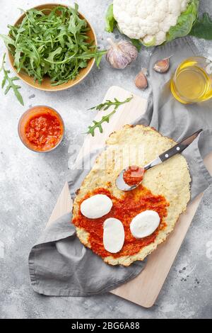 Hausgemachte glutenfreie Cauliflower Pizza Kruste Teig mit Tomaten Marinara-Sauce, Mozzarella-Käse und Rucola auf grauem Hintergrund.Gemüsealternative Stockfoto