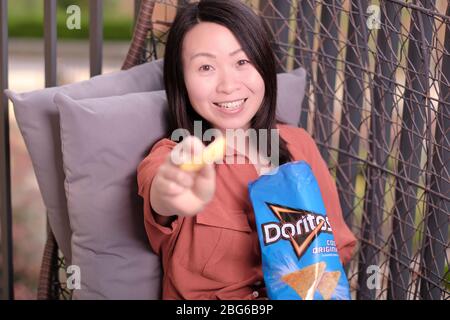 Schöne asiatische Frau auf Rattan Korbsessel auf Balkon sitzen lächelnd und entspannend und bietet einen dorito Chip aus einem blauen Paket von kühlen Original tun Stockfoto