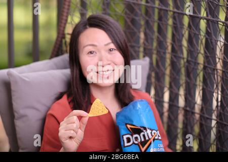 Schöne asiatische Frau auf Rattan Korbsessel auf Balkon sitzen lächelnd und entspannend und bietet einen dorito Chip aus einem blauen Paket von kühlen Original tun Stockfoto