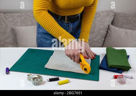 Frau in einem gelben schneidet den Stoff. Es gibt Quilt-Werkzeuge auf dem Tisch. Patchwork-Messer, Schere, gefütterte Schneidematte, selbstschließend, Faden, Zentimeter Stockfoto