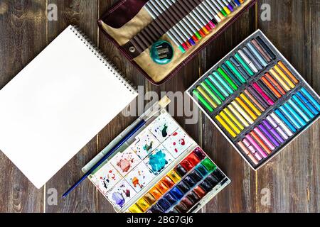 Flacher Kunstarbeitsplatz, Holztisch mit Bleistiften, Pinsel, Aquarellfarben, Papier und Kreiden. Stockfoto