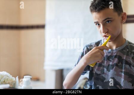 Portrait-Ansicht eines jungen Mannes, der seine Zähne in einem Heim-Badezimmer putzt, auf seine eigene Reflexion schaut, die sich für die Schule vorbereitet, Heim-Interieur. Gesundheit und Stockfoto