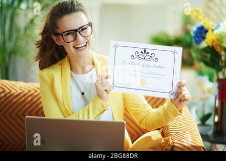Happy stilvolle Frau in Jeans und gelbe Jacke mit Laptop zeigt Abschlusszeugnis in der modernen Wohnzimmer an sonnigen Tag. Stockfoto