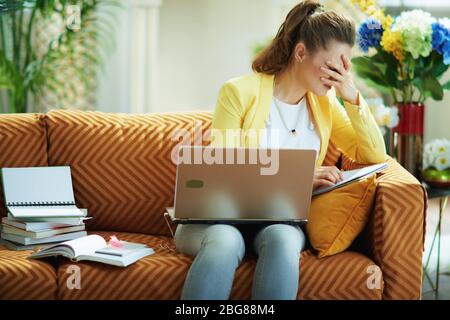 Gestresste moderne Frau in Jeans und gelbe Jacke im modernen Wohnzimmer in sonnigen Tag Studie online auf einem Laptop. Stockfoto