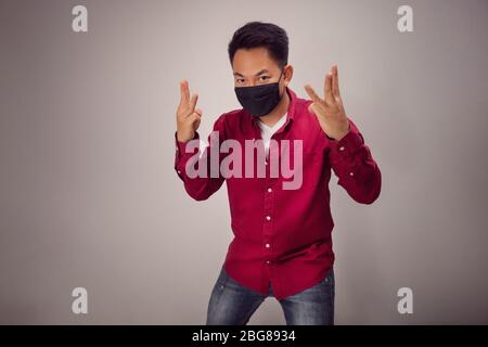 Ein asiatischer Kurzhaarmann in rotem Hemd zeigt in der Studiobeleuchtung mit schwarzer Maske den Finger Nummer drei vor der Kamera. Stockfoto