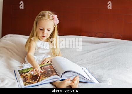 Nettes kleines kaukasisches Mädchen in stilvollem weißen Kleid, das ein Buch liest. Kind auf einem Bett im Zimmer. Bleiben Sie während der Sperrung des Coronavirus Covid-19 zu Hause. Stockfoto