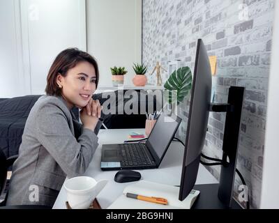 Schöne glückliche asiatische Frau, die mit Laptop-Computer arbeitet und einen anderen Monitor in ihrem Schlafzimmer, Eigentumswohnung aufpasst. Konzept „Arbeit zu Hause“. Stockfoto
