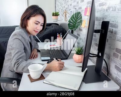 Schöne glückliche asiatische Frau, die mit Laptop-Computer arbeitet und etwas auf Notebook in ihrem Schlafzimmer, Eigentumswohnung schreibt. Konzept „Arbeit zu Hause“. Stockfoto