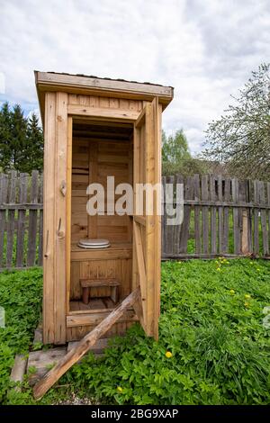 Tradition, rustikale HolzToilette, WC, mit einem modernen Toilettensitz mit Deckel, im Hof, vor dem Hintergrund eines alten Zauns. Stockfoto