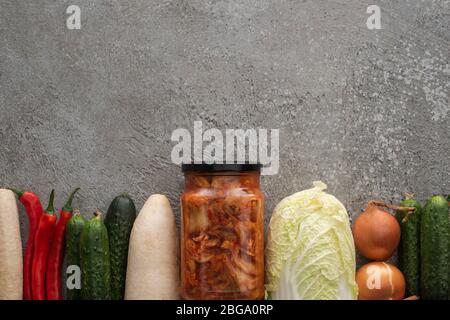 Draufsicht von Chilischoten, Gurken, Daikon Rettich, chinakohl, Zwiebeln und Kimchi-Glas auf grauem Beton Hintergrund Stockfoto