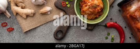 Panorama-Schuss Kimchi in Schüssel und Glas auf Schneidebrett in der Nähe von Ingwer, Knoblauch und Chili Pfeffer auf Betonoberfläche Stockfoto