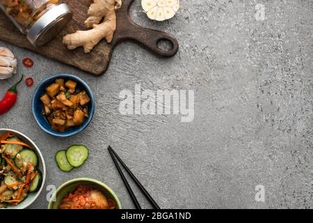 Draufsicht von leckeren Kimchi in Schüsseln und Glas in der Nähe Schneidebrett, Essstäbchen, Knoblauch, Ingwer und Chili auf Betonoberfläche Stockfoto