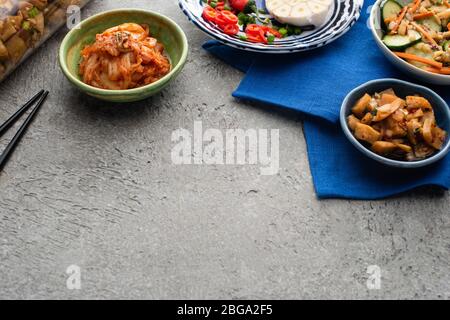 Leckere Kimchi in Schüsseln und Glas auf blauem Tuch in der Nähe von Essstäbchen auf Betonoberfläche Stockfoto