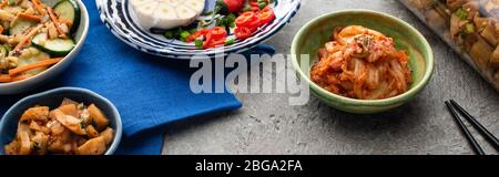 Panorama-Aufnahme von leckeren Kimchi in Schüsseln und Glas auf blauem Tuch in der Nähe von Essstäbchen auf Betonoberfläche Stockfoto