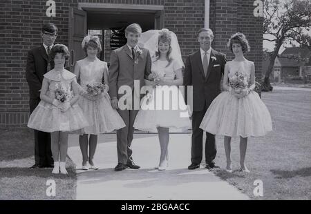 Historisches Gruppenbild der 1960er Jahre, das Hochzeitsgäste mit einer "Teenager"-Braut in ihrem Brautkleid mit Brautschleier und Bräutigam in einem Dreiknopf-Anzug der damaligen Zeit zeigt, die vor dem Eingang eines modernen Kirchengebäudes in England zusammenstehen. Stockfoto