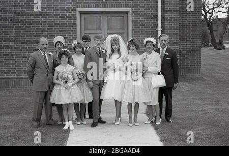 Historisches Gruppenbild der 1960er Jahre, das Hochzeitsgäste mit einer "Teenager"-Braut in ihrem Brautkleid mit Brautschleier und Bräutigam in einem Dreiknopf-Anzug der damaligen Zeit zeigt, die vor dem Eingang eines modernen Kirchengebäudes in England zusammenstehen. Stockfoto