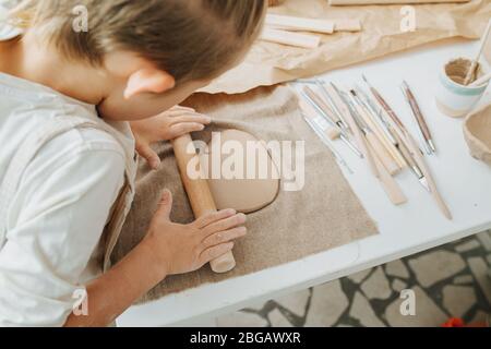 Boy rollenden Ton Stück auf einem Tisch mit Töpferei-Werkzeuge gefüllt. Stockfoto