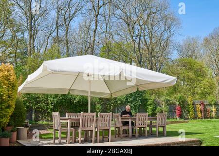 Der ältere Mann sitzt an einem sonnigen Frühlingstag unter einem Sonnenschirm auf einer Terrasse im Garten. Stockfoto