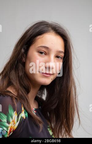 Attraktive junge Frau mit einem ruhigen Lächeln und langen braunen Haaren drehen, um nachdenklich auf die Kamera über einen grauen Hintergrund zu schauen Stockfoto