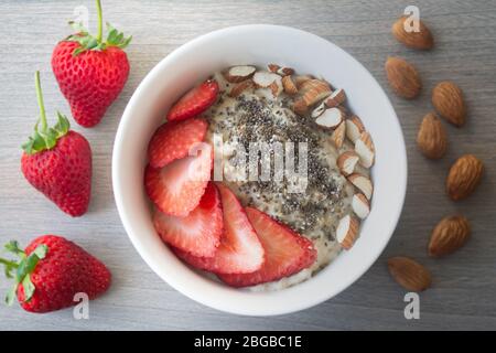 Gesunde vegane Haferflocken mit Erdbeeren, Chiasamen und in Scheiben geschnittenen Mandeln in einer weißen Schüssel mit Erdbeeren und ganzen Mandeln im Hintergrund Stockfoto