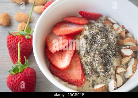 Gesunde vegane Haferflocken mit Erdbeeren, Chiasamen und in Scheiben geschnittenen Mandeln in einer weißen Schüssel mit Erdbeeren und ganzen Mandeln im Hintergrund Stockfoto