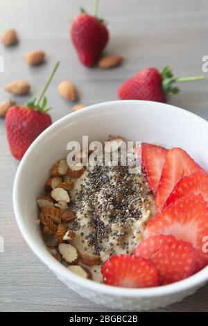 Gesunde vegane Haferflocken mit Erdbeeren, Chiasamen und in Scheiben geschnittenen Mandeln in einer weißen Schüssel mit Erdbeeren und ganzen Mandeln im Hintergrund Stockfoto